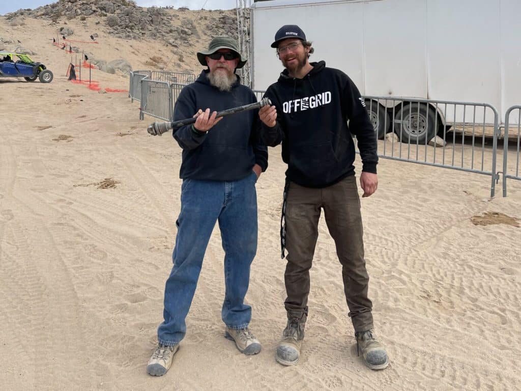 Two men stand side-by-side holding broken truck parts.