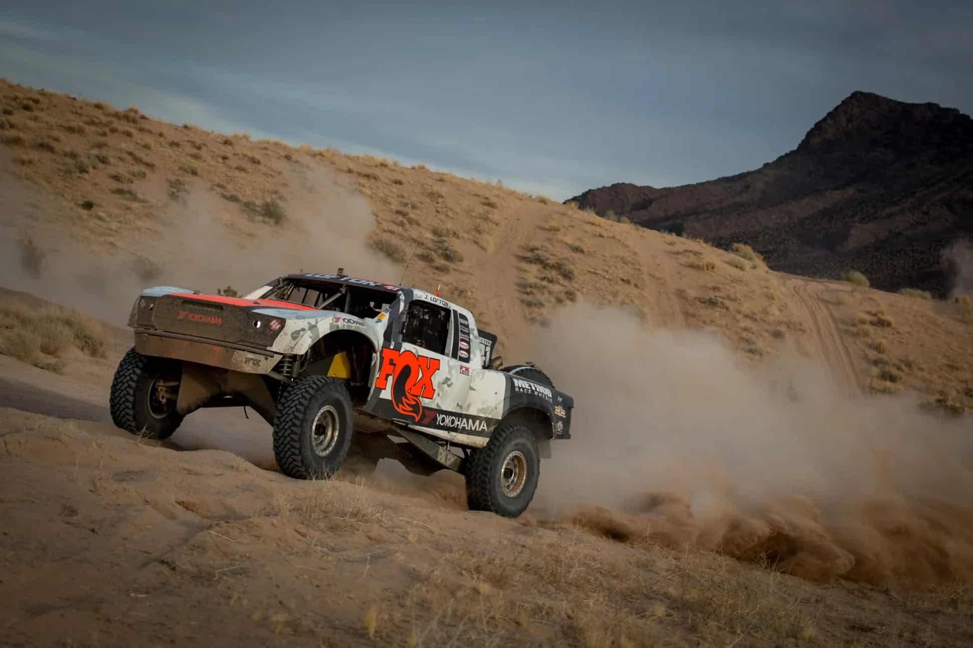 A racing truck driving across dirt terrain throwing dust behind it.