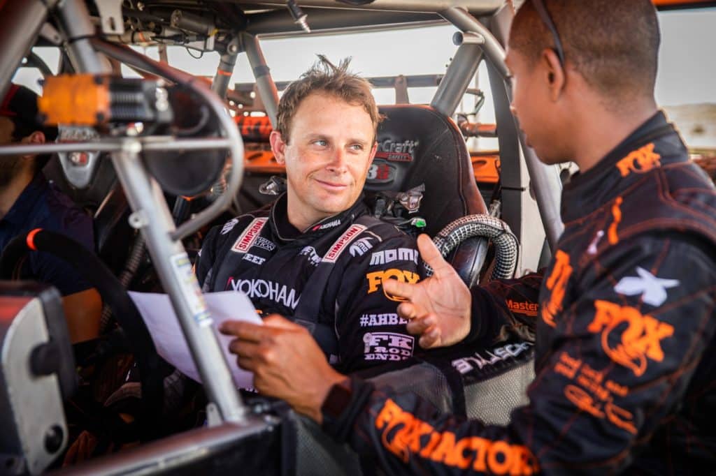 A man sits in a racing truck cab talking to another man.