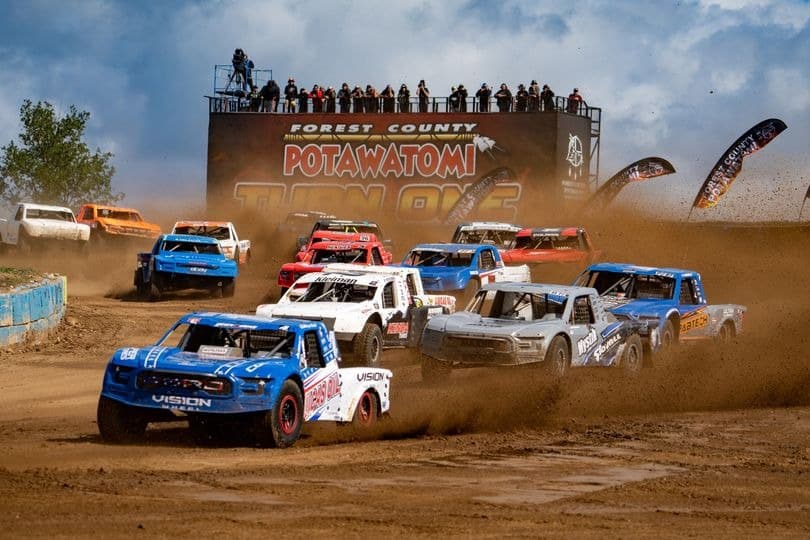 Trucks race around the turn of a dirt track, spraying dirt and mud through the air.