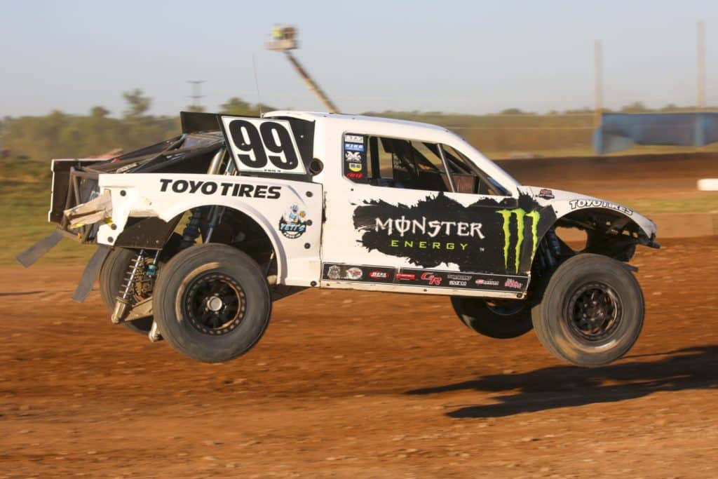 A racing truck leaping through the air on a dirt racing track.