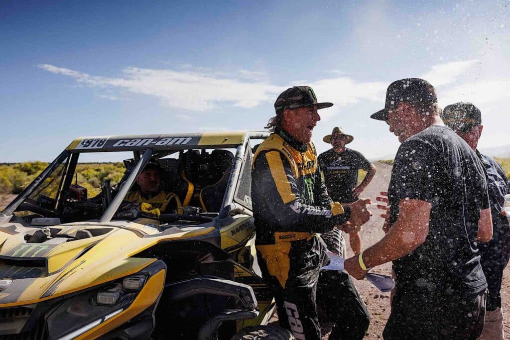 A group of people standing around a Can-Am in the desert.